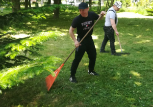 Chłopak ubrany na czarno i drugi w jasnej podkoszulce grabią liscie w ogórdku szkolnym.